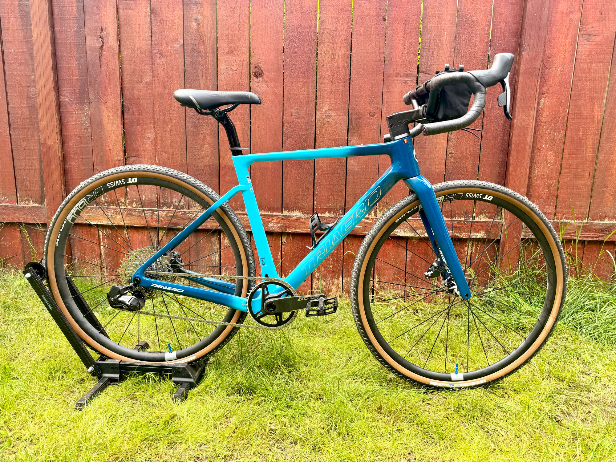 A teal Triaero Grava gravel bike on a workshop stand against a red cedar fence — DT Swiss alloy wheels with tan-wall tires and a Redshift raised handlebar.