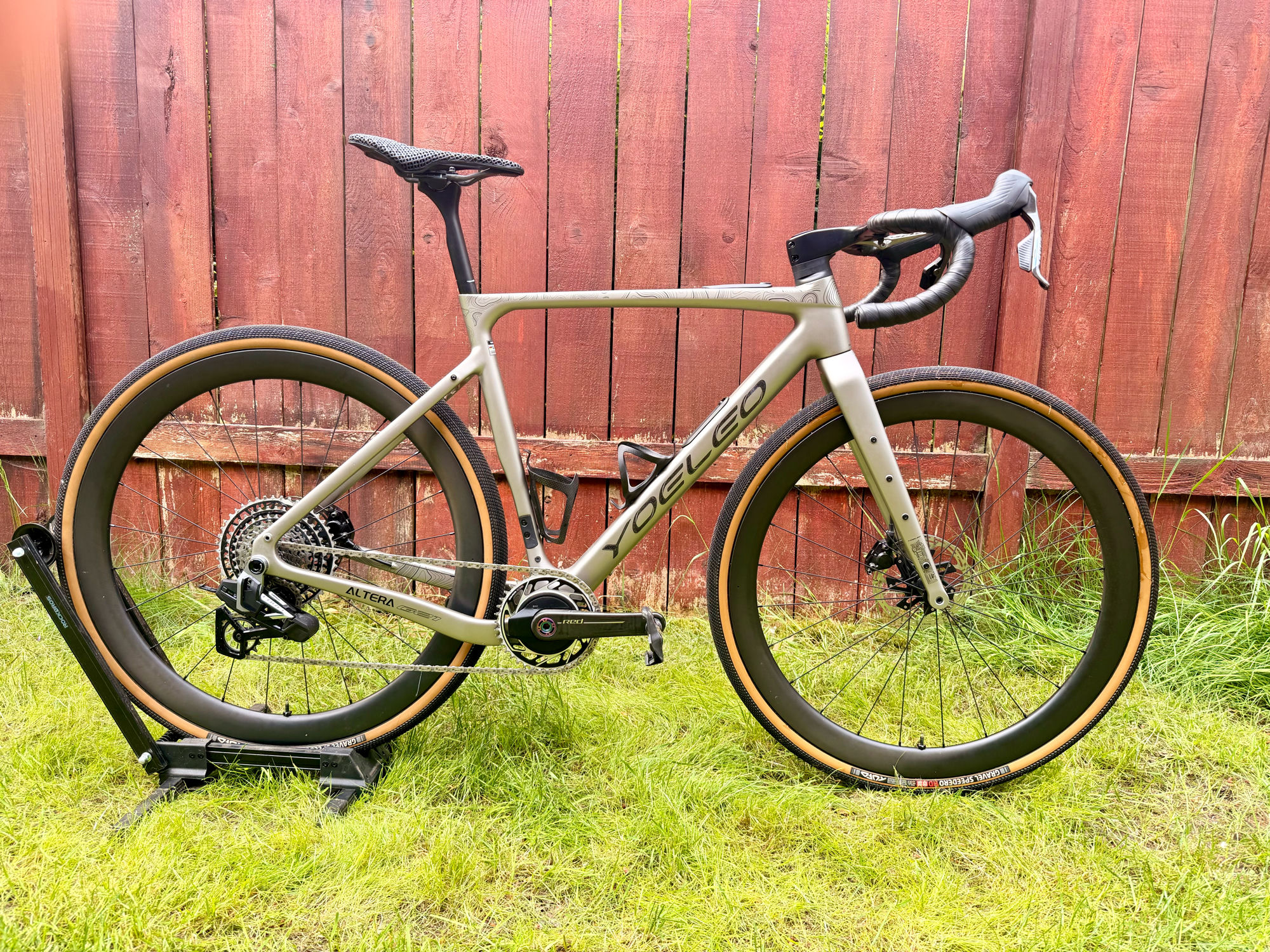 A champagne Yoeleo Altera G21 gravel frame on a workshop stand against a red cedar fence — SAT C45 carbon wheels with tan-wall Speedero tires and SRAM Force XPLR E1 with a Quarq spider power meter.
